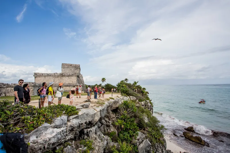 The Tulum ruins: a Mayan temple of gray stone on the cliff above the Caribbean Sea, with visitors on the path and a small boat offshore
